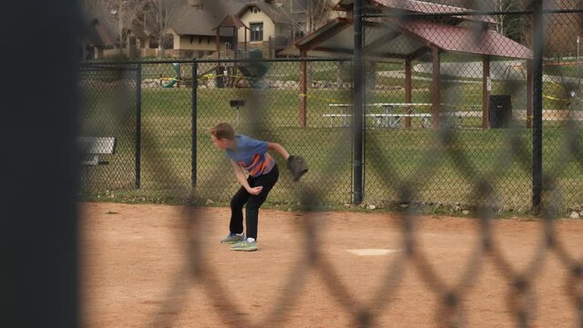 Behind the fence shot of the young baseball player. Boy is training to catch the ball and then throws it.