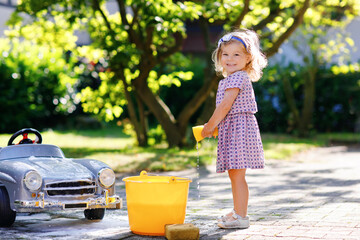 Cute gorgeous toddler girl washing big old toy car in summer garden, outdoors. Happy healthy little...