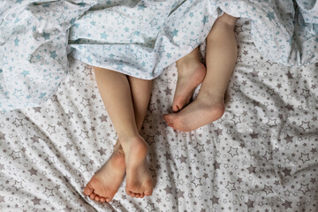 Close-up of two pairs of toddler girls feet on the bed under the blanket. Light blue and beige tones.