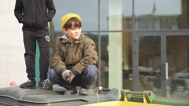 Lonely street children in dirty wear spend time together outdoors, sitting on trash cans, rubbish.