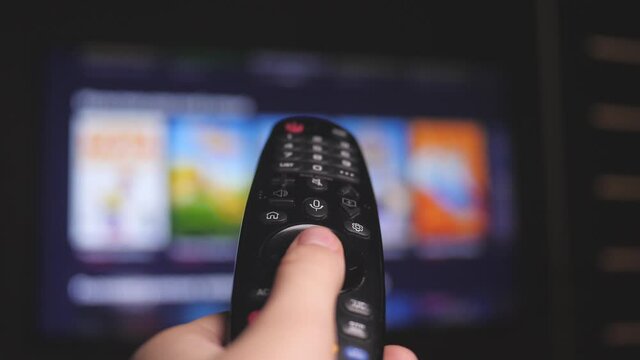 Woman hand selects internet tv channels with remote control, close-up. Person controls TV using a modern remote control. Girl watches smart TV and uses black remote control. Blurry tv scrolls pages