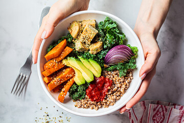Woman hands eating vegan salad of baked vegetables, avocado, tofu and buckwheat buddha bowl, top view. Plant based food concept. - obrazy, fototapety, plakaty