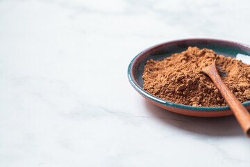 Raw organic cocoa powder on saucer with wooden spoon, white marble background. Super food concept.