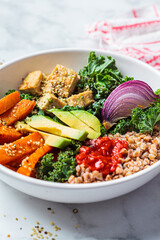 Baked vegetables, avocado, tofu and buckwheat buddha bowl. Vegan lunch salad with kale, baked sweet potato, tofu, buckwheat and avocado in white bowl. Vegan food concept.
