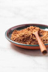 Raw organic cocoa powder on saucer with wooden spoon, white marble background. Super food concept.
