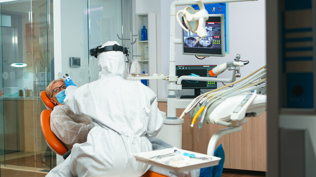 Nusre with face shield measuring old woman temperature before dental examination during global pandemic. Concept of new normal dentist visit in coronavirus outbreak wearing protective suit and face