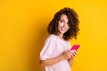 Photo portrait of curly curious girl keeping smartphone looking at blank space isolated on vivid yellow color background