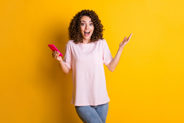 Photo portrait of shocked woman with open mouth holding phone in one hand isolated on vivid yellow colored background