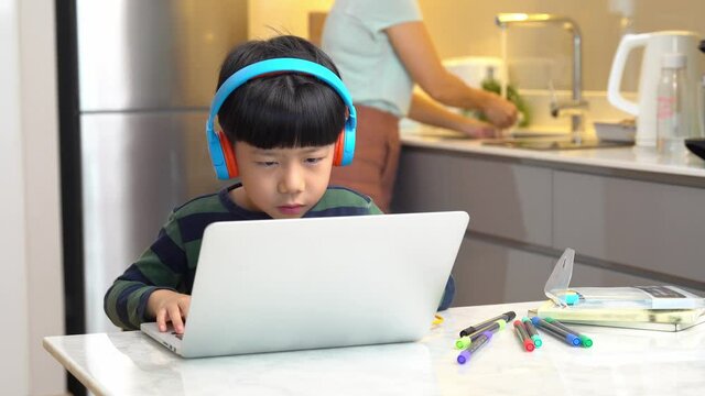 A smart asian little boy learning online at home during Covid-19 pandemic, he use computer laptop to join the virtual classroom from school while his mother doing housework in the kitchen. New normal.