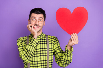 Photo of young excited shocked amazed happy positive man bite fingers hold big red heart isolated on violet color background