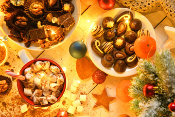 Fototapeta na wymiar sweet food top view background for merry christmas or new year holiday decoration with night illumination - chocolate candies, tangerines, cookies, marshmallow and cocoa latte on white wood