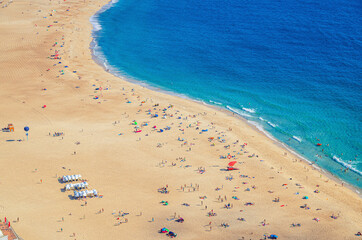 Top aerial view of sandy beach with people tourists sunbathing and Atlantic Ocean azure blue water, Praia da Nazare town coastline with waves, Leiria District, Oeste region, Portugal