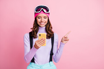 Photo portrait of smiling woman pointing finger at blank space holding phone in one hand isolated on pastel pink colored background