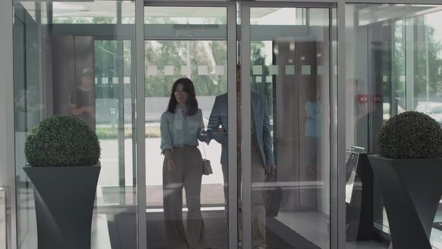 Medium shot of young woman and middle-aged man in elegant outfits walking together into car dealership through sliding glass doors