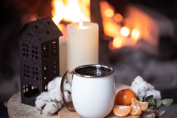 Cozy composition with a cup, candles and tangerines on a blurred background of a burning fireplace.