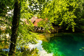 Scenic view of the natural river head called Blautopf in Blaubeuren, Germany. A historic hammer mill reflects in the blue-turquoise water.