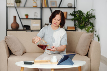 Elegant senior woman sitting on sofa at home and pouring herself a cup of coffee