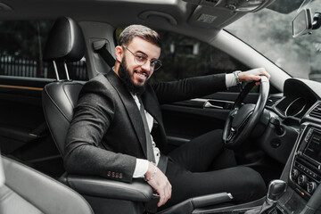 Obraz premium Portrait of a handsome elegant man in a suit sitting behind the wheel and looking at the camera with a smile.