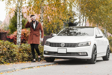 Stylishly dressed young man walking past his expensive car. Successful young man walks along the autumn street with a smile