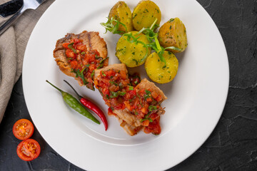 stewed pieces of meat with spices. With baked new potatoes and tomato salsa sauce on a white platter and dark background. Dish for the restaurant menu