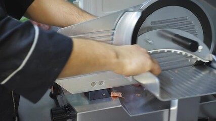 Arm of cook using cutting machine for salami at cuisine restaurant. Male hand of chef chopping sausage on electric food slicer at kitchen. Process to prepare ingredients for cooking pizza. Close up