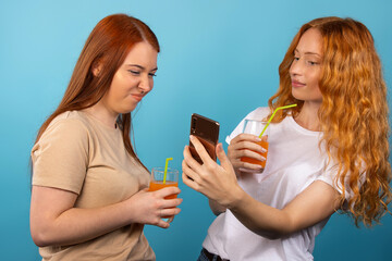 Young girls take a selfie, drink multifruit juice from a straw and look at photos on a smartphone.