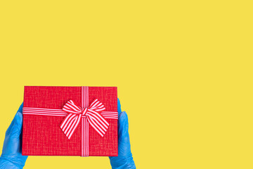 Safe delivery of gifts concept. Hands in blue gloves hold a red gift box with a bow on a trendy Illuminating yellow background. Safe gifts during the coronavirus pandemic