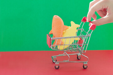 A hand holds a shopping cart on a red and green background with plastic fish inside. The concept of...