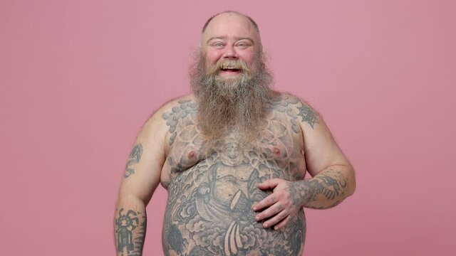 Laughing funny fat pudge obese chubby overweight bearded man has tattooed naked bare big belly isolated on pastel pink background studio. People sincere emotions lifestyle concept. Looking at camera