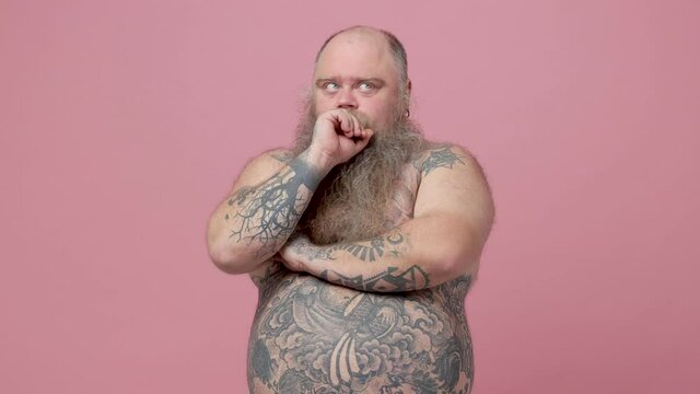 Pensive fat pudge obese chubby overweight bearded man has tattooed naked bare big belly isolated on pastel pink background studio. People lifestyle concept. Looking aside up put hand prop up on chin