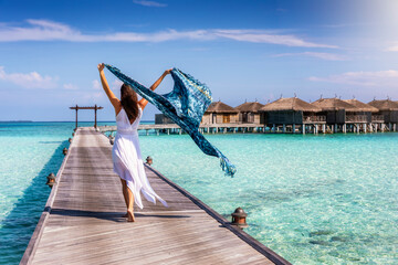 A beautiful tourist woman walks on a wooden pier over turquoise ocean in the Maldives and holds a waving scarf in her hands