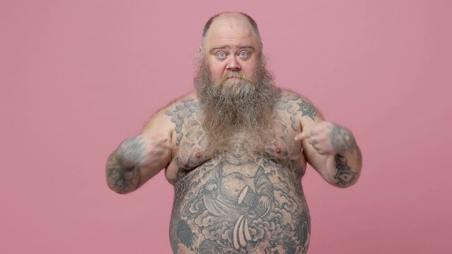 Shocked fat pudge obese chubby overweight bearded man has tattooed naked bare big belly isolated on pink background. People lifestyle concept. Point index fingers thumbs on himself hold hands crossed