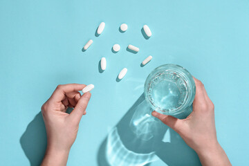 Woman holding in hands white medical pill and glass of water on light blue background, copy space. Top view, fat lay. Medicine concept. Pharmaceutical - obrazy, fototapety, plakaty
