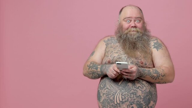 Shocked fat pudge obese chubby overweight bearded man has tattooed naked bare big belly isolated on pink background studio. People lifestyle concept. Using mobile cell phone point index finger aside