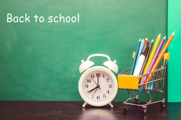 White alarm clock, a shopping cart with colored pencils stands on the table against the background of a green blackboard with the text Back to school. Concept of school, study, education.