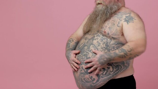 Cheerful fat pudge obese chubby overweight bearded man stroking shaking touching tattooed naked bare big belly isolated on pastel pink background studio. People sincere emotions lifestyle concept