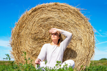 Naklejka na ściany i meble Sunny day. A woman in white clothes sits by a haystack on a mown field. She looks into the distance, covers her eyes from the sunlight with her hand.