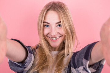 Portrait of a happy young girl with straight hair, in a shirt, taking a selfie on a pink background