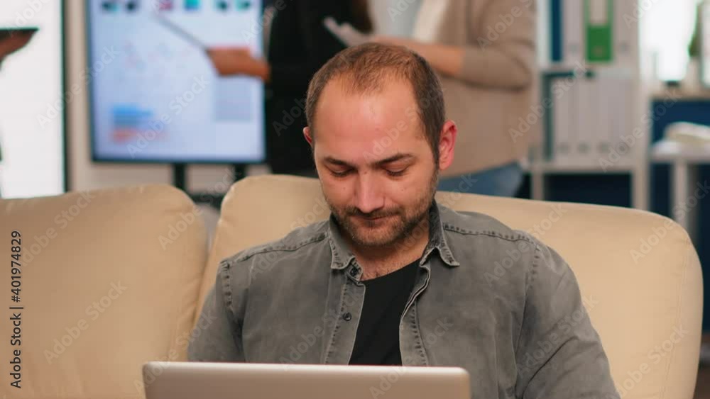 Poster manager sitting on couch in front of camera smiling while multiethnic team working in background wit