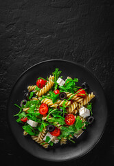 Salad with pasta, arugula, tomato, cheese and olives on black plate on Dark grey black slate background