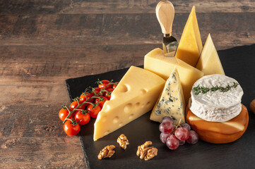 Collection of Swiss, French, Italian and Holland cheeses on a black cheese board served with fruit, nuts on black slate board on rustic wooden table