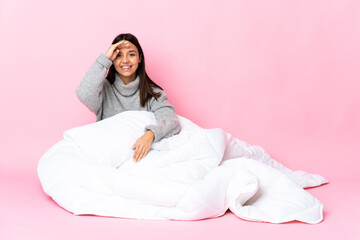 Young mixed race woman wearing pijama sitting on the floor saluting with hand with happy expression