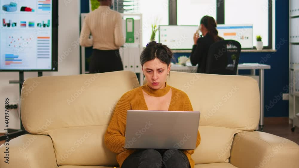 Poster concentrated manager reading mails typing on laptop sitting on couch in busy start up office while d