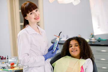 Child at dentist's office. Little patient, mixed raced curly girl, sitting in dentistry chair, looking at camera and smiling. Pretty female dentist posing to camera with UV polymerization lamp