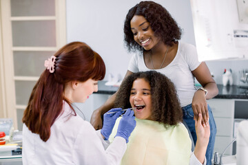 Little mixed raced school girl with curly hair, visiting dentist for checkup or caries treatment, sitting in dental chair and holding hand of her pretty African mom. Female dentist makes examination.