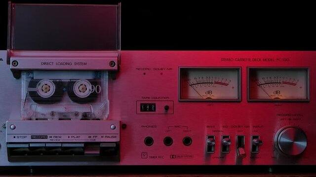 Vintage cassette player deck from 80s illuminated by red neon lights. Stereo recorder with rotating cassette, controls, dashboard and buttons on black studio background. Close up. Slow motion.