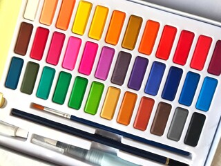 A palette of bright multicolored watercolors, brush and pencil close-up, artist's tools all colors of the rainbow, selective focus of paint