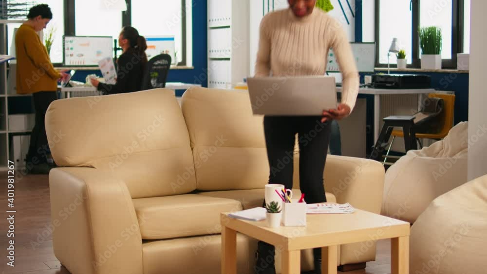 Poster focused black manager reading tasks on laptop and typing sitting on couch in busy start up office wh
