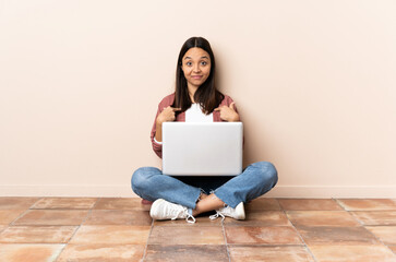 Young mixed race woman with a laptop sitting on the floor with surprise facial expression