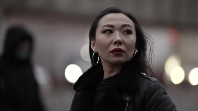 Close-up of a beautiful and spectacular Asian woman with long dark hair in a stylish image, she walks on a city street against the background of evening lights. The people passing by.